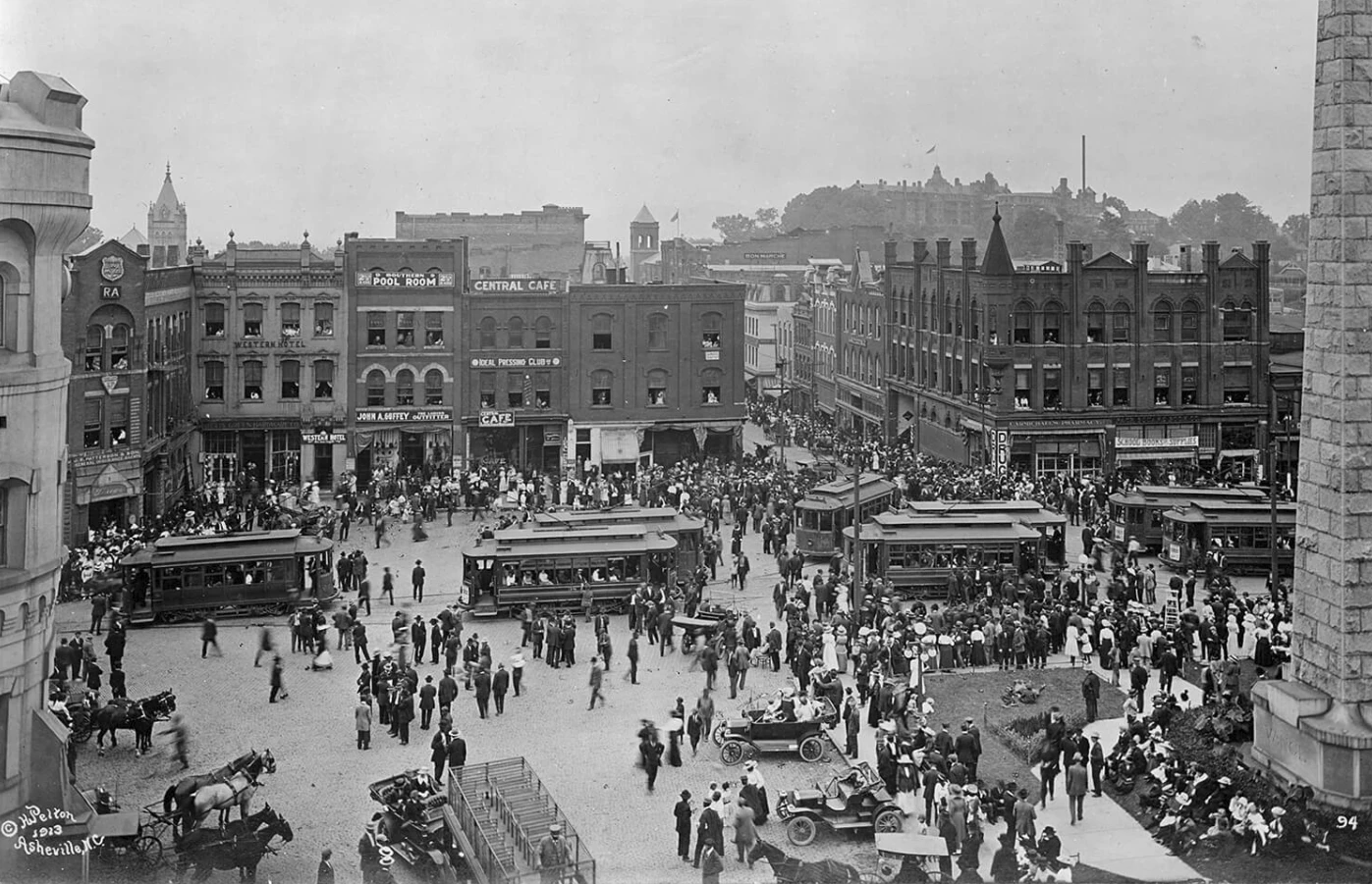 Black and white history photo of Asheville, NC from the 1920's showing busy Pack Square filled with people walking and numerous streetcars. Surrounding the square are multi-story brick buildings with shops. The street is bustling with activity, reflecting early 20th-century urban life.