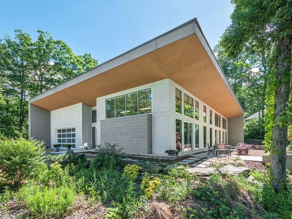 A modern house with large glass windows and a slanted roof, surrounded by greenery and trees. The architecture features clean lines and a mix of concrete and wood materials in a serene natural setting under a clear blue sky.
