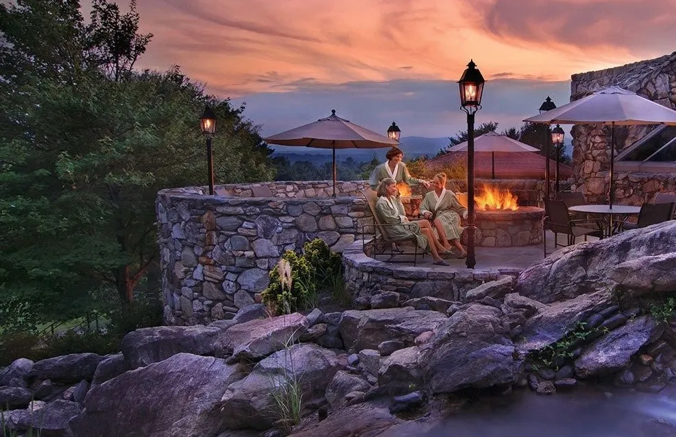 Three people in robes sit by a fire pit on a stone patio at sunset. They are surrounded by lanterns and trees, with tables and umbrellas nearby. The scene overlooks a scenic landscape with a colorful sky.