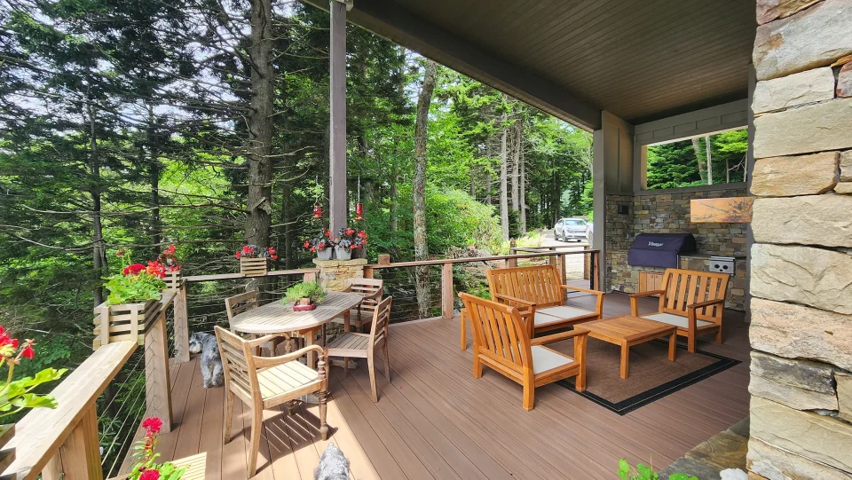 A wooden deck in a forest setting features a dining table with chairs and a seating area with benches. Flower pots with red blooms adorn the railing. A grill is visible in the corner. Tall trees surround the deck, offering a serene, natural ambiance.