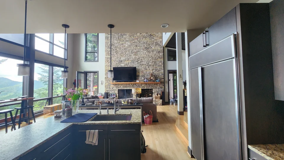 Spacious modern kitchen and living area with high ceilings and large windows. Features dark cabinetry, a central island with a sink, a stone fireplace, and views of greenery outside. Cozy and well-lit with pendant lights.