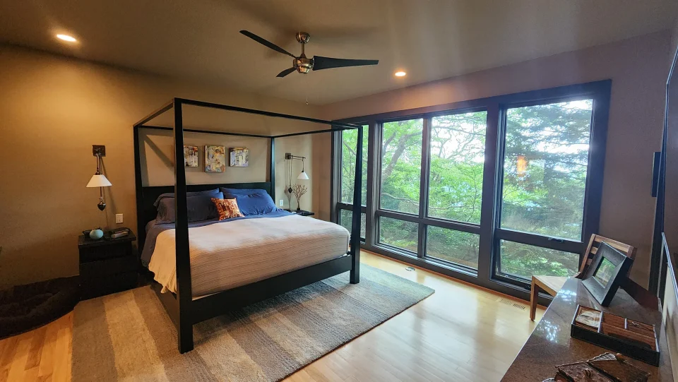 Photo of a bedroom with a modern ceiling fan, modern furniture a wall of windows with trees outside.