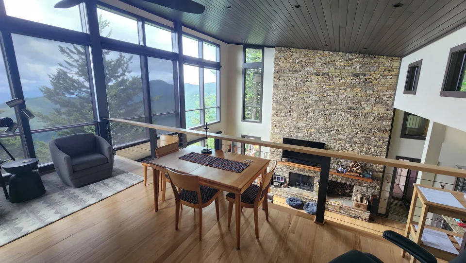 Photo of an interior balcony on a second floor overlooking a large living room with a vaulted ceiling and a wall of windows overlooking a beautiful mountain vista.