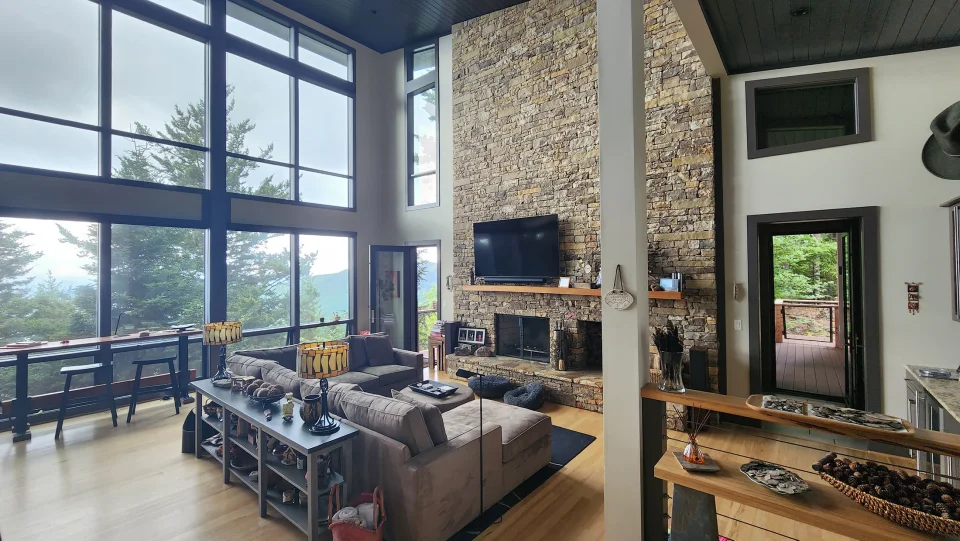 Photo of a modern living room with a stone wall with a fireplace, vaulted ceiling, one wall made of windows, a couch and door in the background opening up to a deck.