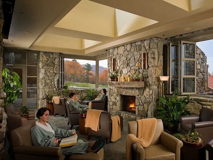 People relaxing in a cozy lounge area with stone walls, large windows, and a fireplace. They are seated in armchairs, wearing robes. The room is decorated with plants, and outside the windows, fall foliage is visible.
