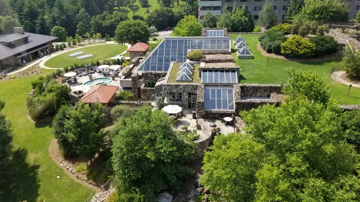 Aerial view of a large, modern building with glass roofs surrounded by lush greenery. There's a pool area with loungers on one side and a large garden with trees and pathways. A few buildings are visible in the background.
