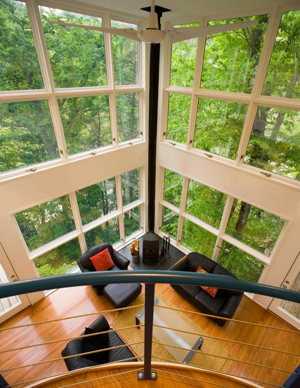 A modern two-story living room with floor-to-ceiling windows displaying a lush forest. The space has a black sofa set, a coffee table, and a central black stove, viewed from a mezzanine balcony with a curved metal railing.