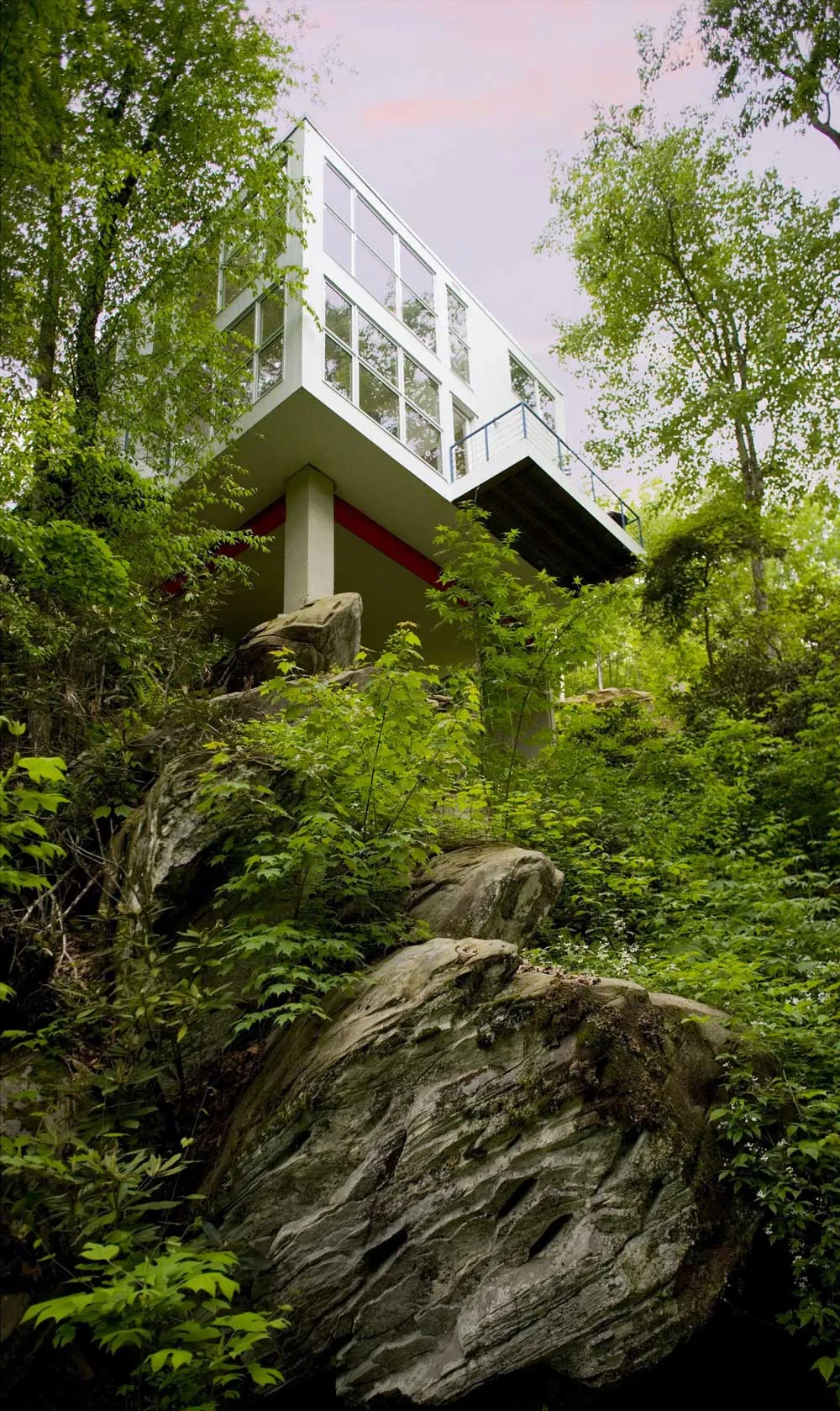 A modern, white rectangular house with large windows is perched on stilts, surrounded by lush green trees and vegetation. It's built over rocky terrain, blending into the natural landscape under a pinkish sky.
