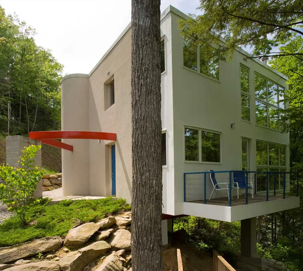 Modern two-story house with large windows, a red curved awning, and a blue railing. Surrounded by trees and set on a rocky hillside, the home has a patio with chairs and blends into the lush greenery.