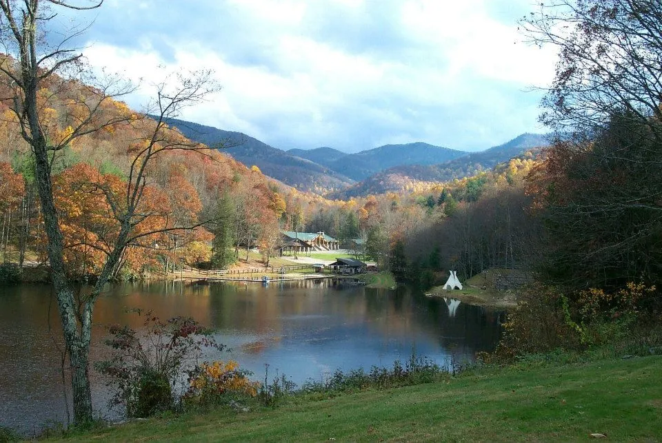 A scenic view of a lake reflecting surrounding autumn trees with colorful foliage. A house and a teepee are visible by the lakeside. The backdrop features rolling mountains under a partly cloudy sky.