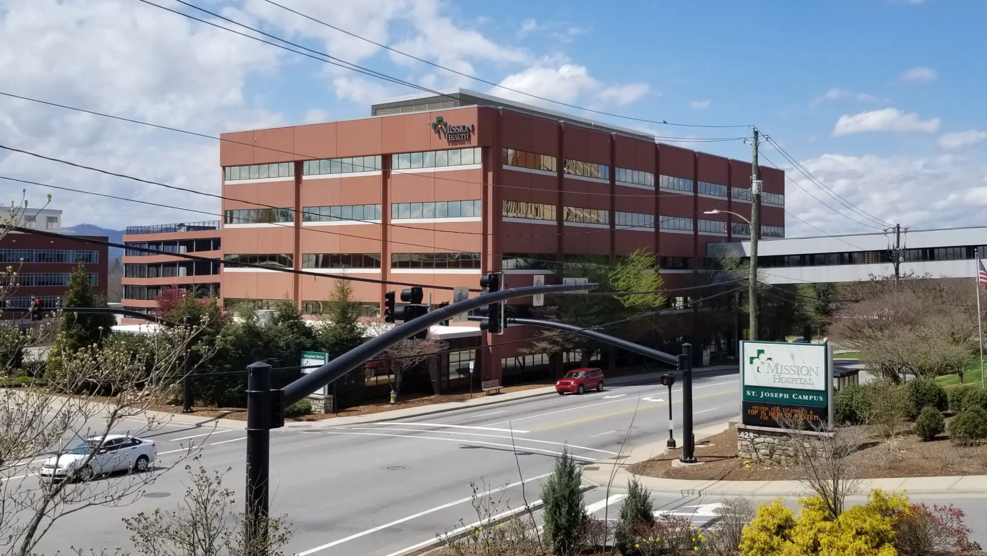 A large brick building with a sign for Mission Health is visible on a sunny day. In the foreground, a traffic light and street intersection with a sign for St. Joseph Campus can be seen. Trees and a blue sky with clouds are in the background.