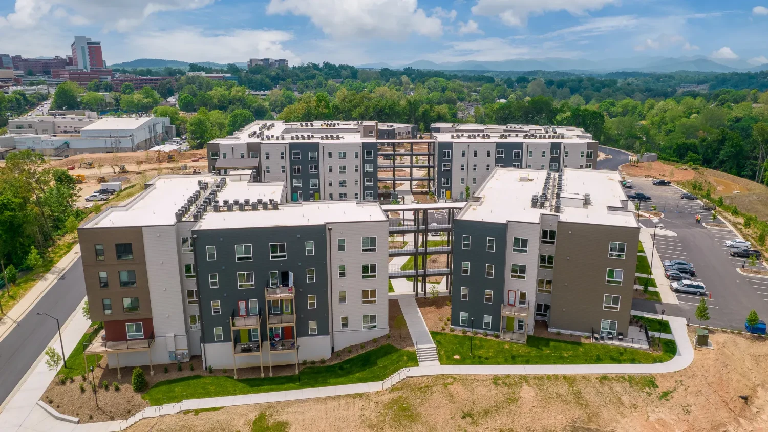 Aerial view of a modern apartment complex with multiple buildings. Each building has a mix of dark and light gray exteriors. Surrounding the complex are green trees, a parking area, and a road. A cityscape is visible in the background under a partly cloudy sky.