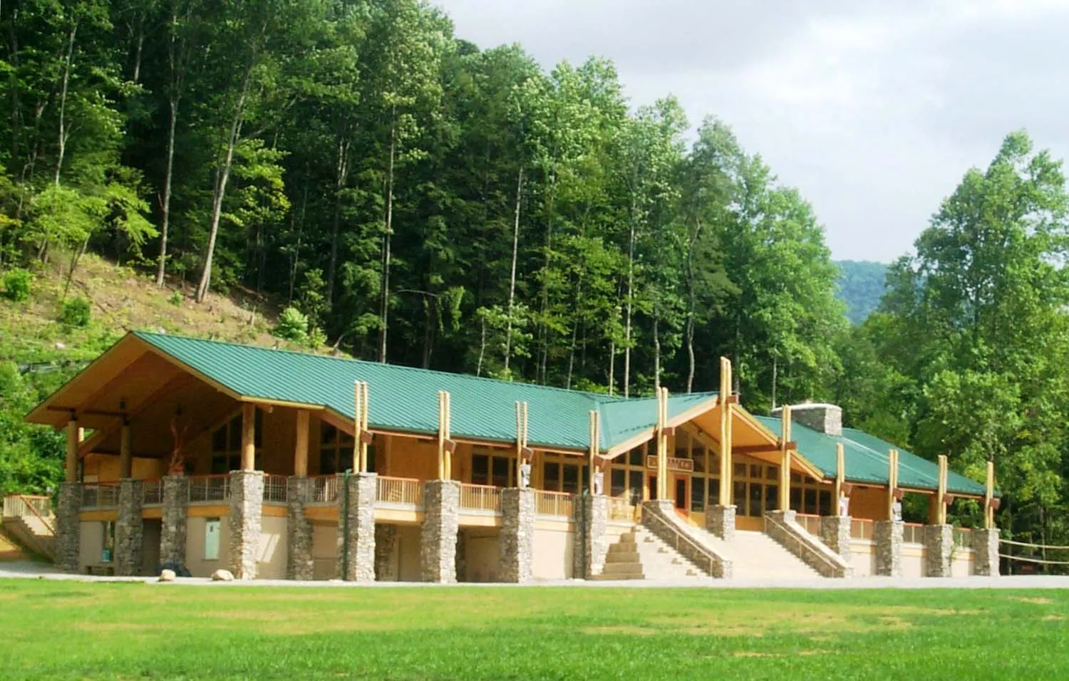 A large wooden lodge with a green roof is set against a backdrop of tall trees and hills. The building features stone pillars and twin staircases leading to the entrance. The sky is partly cloudy, and the surrounding area is grassy.