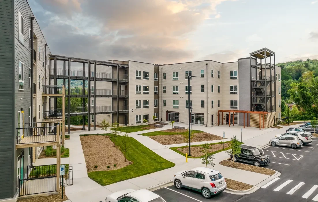 Modern apartment complex with multiple stories and balconies. Landscaped courtyard with small trees and shrubs. Several parked cars are in the adjacent parking lot. The sky above is partly cloudy.