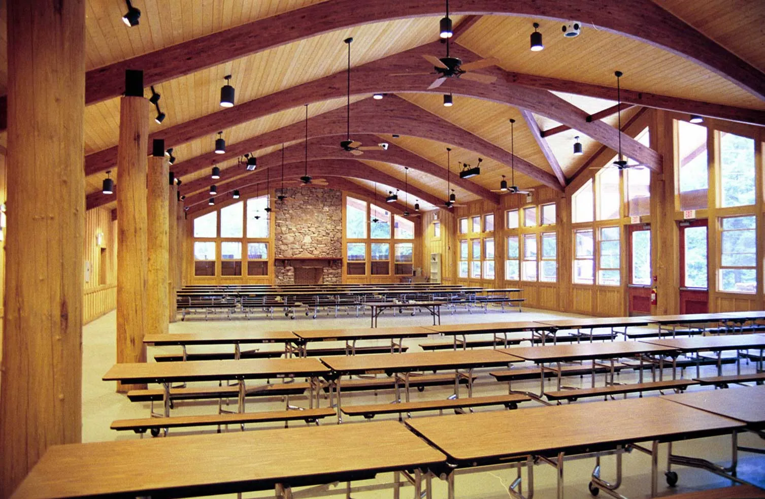 A spacious dining hall with wooden beams and a high ceiling. The room features multiple long tables and benches, a stone fireplace at the far end, and large windows along the walls, allowing natural light to brighten the space.