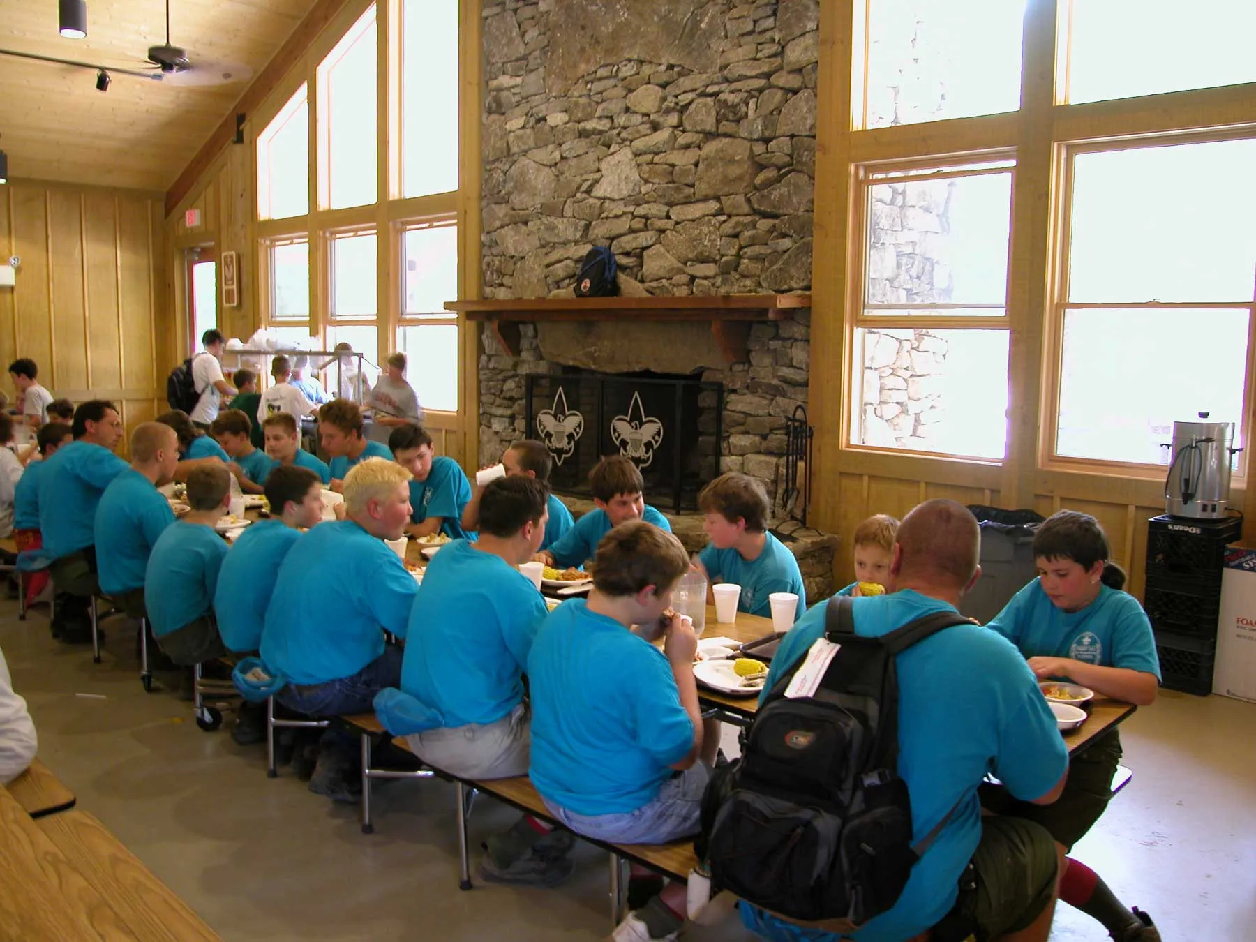 A group of people wearing matching blue shirts sit on benches around long tables in a rustic dining hall. They are eating and talking. A stone fireplace with scout emblems is visible in the background, and large windows let in natural light.