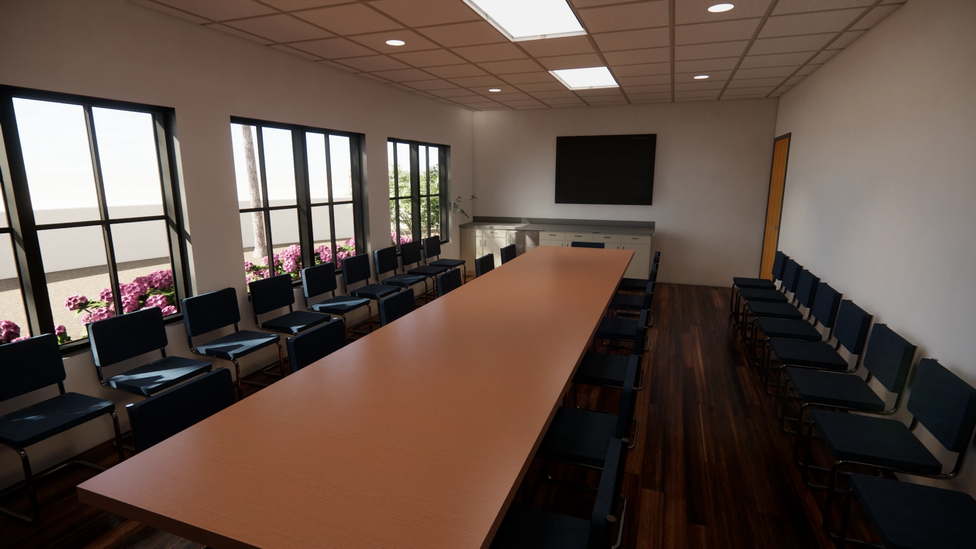 A long, empty conference room with a rectangular wooden table surrounded by chairs. Large windows line the left wall, letting in natural light, and there is a black screen on the wall at the far end of the room.
